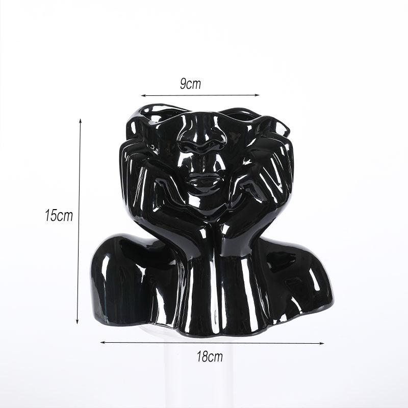 Glossy black ceramic face vase with hands framing the face, shown 15 cm high and 18 cm wide