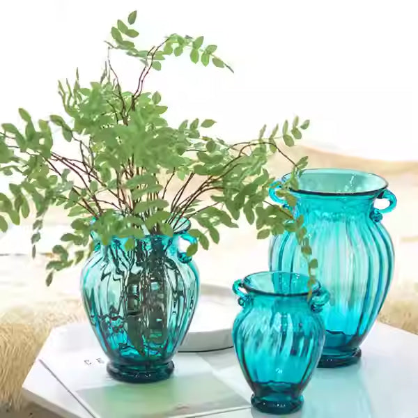 Three teal ribbed glass vases with twin handles holding greenery